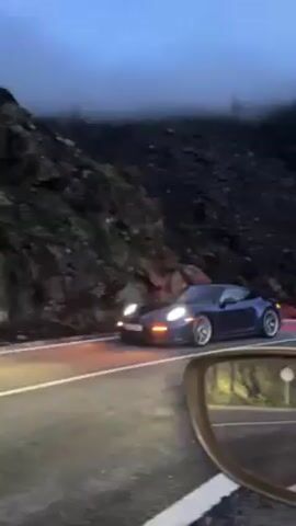 The video shows a blue Porsche driving on a wet mountain road. The road is bordered by a steep, dark rock face on one side. Fog hangs in the air, obscuring the upper part of the mountain. The camera appears to be in another car, as a side mirror is visible in the lower right of the frame. The Porsche's headlights and taillights are illuminated. The car is moving forward along the road.