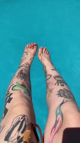 The video shows a pair of legs submerged in bright blue water. The legs have multiple tattoos covering them, including what appears to be a checkered pattern, abstract designs, flowers, and a flame-like shape. The feet are visible and are also in the water. Small ripples are visible on the surface of the water around the feet. The camera angle is from above, looking down at the legs and water. A brown object, possibly part of a bag or accessory, is visible in the lower right corner of the frame. The lighting suggests it is daytime and sunny.