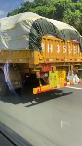 The video shows a close-up, side view of a yellow semi-trailer truck traveling on a road. The truck has a large tarp covering its cargo. The tarp is white with yellow stripes and blue and red stripes near the bottom. The truck's wheels and mud flaps are visible, as well as its side marker lights. There are hazard symbols on the side of the trailer. A white semi-truck cab is partially visible on the right side of the frame. The environment appears to be a sunny day with trees and a blue sky visible in the background. The camera is moving, suggesting the video was recorded from another vehicle.