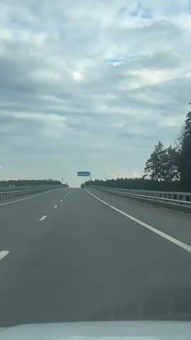 The media file depicts a view from a vehicle traveling on a highway. The highway has multiple lanes, marked with white lines, and is bordered by metal guardrails. On the right side of the highway, a dense line of trees forms a forest. The sky is overcast with dramatic, gray clouds, with a few patches of blue sky visible. There is a blue highway sign in the distance, which appears to have text and a parking symbol on it. The scene suggests a daytime journey through a rural or semi-rural area. The weather appears to be cloudy, possibly before or after rain. The specific location is noted as Demikhovo, Russia. No people are clearly visible in the frame. The overall mood is one of travel and transition.