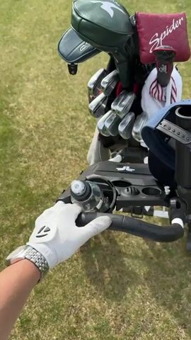 The video shows a person's hand, wearing a white golf glove and a silver wristwatch, reaching for a clear water bottle nestled in the holder of a black golf cart. The cart is laden with a full golf bag containing an assortment of clubs with various headcovers, including a dark green one, and a red one labeled "Spider". A white towel with red stripes and a blue baseball cap are also attached to the cart. The cart stands on a grassy lawn, presumably part of a golf course.