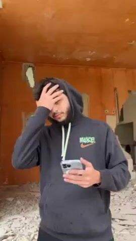 A young man with dark hair and a beard stands in what appears to be an unfinished or abandoned building. He is wearing a dark gray Nike hoodie with light yellow drawstrings. He is holding a silver smartphone in his hands, initially holding his hand to his forehead as if frustrated. He then looks at his phone, examining the screen and showing it to the camera. The building behind him has orange walls, and there is debris on the ground.