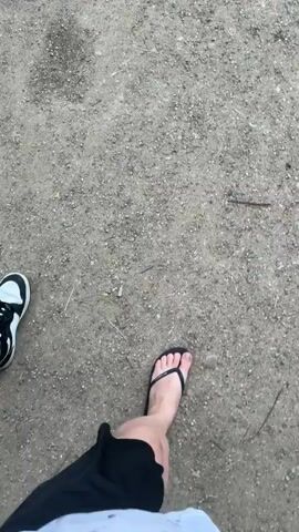 Two people are walking on a gravel path. One person is wearing black sneakers and ripped jeans. The other person is wearing black flip-flops and black shorts. The camera is at a low angle, looking down at their feet. The video appears to be a point-of-view shot.