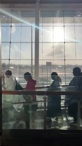 The video shows people walking on a ramp in an airport terminal. The ramp has glass walls and a wooden railing. Outside the window, there is an airport tarmac with planes and a sunny sky with clouds. Several people are seen walking on the ramp, some individually and some in groups. One person in the foreground is wearing a red hoodie and carrying a bag. Another person is wearing a dark jacket. A third person is wearing a light-colored shirt and dark pants. The sun is shining brightly through the window, creating lens flare. The overall atmosphere appears to be that of a busy airport during the day.