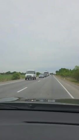 The video shows a car driving down a highway on a cloudy day. In front of the vehicle, there is a car transport truck hauling vehicles. The passenger of the car films the truck, and then the video cuts to a view of the green farmland alongside the highway.