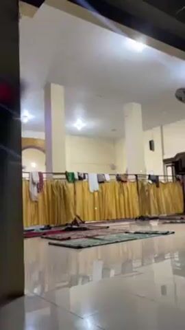 The camera peers into a mosque. In the foreground, the shiny tiled floor reflects the room's interior. Multiple green and red prayer rugs are laid out on the floor, presumably for worshipers. A long golden curtain is hung on a railing, acting as a partition. A variety of clothes, including white and colored pieces, are hanging over the railing, atop the curtain. The mosque has tall cream-colored pillars and walls. Several ceiling lights illuminate the space. The camera is positioned outside the main hall, viewing the scene through an opening.