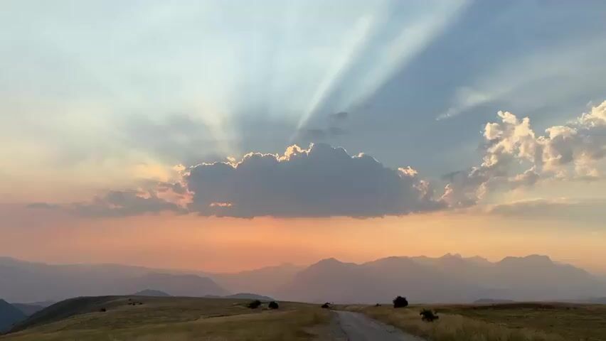 A paved road runs through a grassy, hilly landscape. The sky is filled with dramatic crepuscular rays emanating from behind a large cloud. The sun is low on the horizon, casting a warm, orange glow on the distant mountains. The grass is dry and golden, suggesting a late summer or early autumn setting. No people are clearly visible in the foreground.