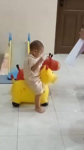A toddler sits on a yellow inflatable toy animal that resembles a horse or cow. The child is wearing a light brown short-sleeved shirt and shorts. The toy has two horns on its head and red ears. In the background, another similar inflatable toy with a red head is visible. To the left, a portion of a children's slide is visible. A woman in a pink headscarf and matching pink outfit stands to the right, facing away from the child, holding a white piece of paper or fabric. The floor is tiled. The wall behind the child is a pale cream color, and there is a wooden door in the background to the right. The overall scene suggests an indoor play area or living space.