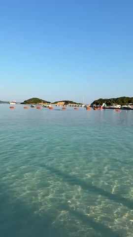 This is a vertical, slightly shaky, exterior shot of a clear blue sky over a calm, turquoise body of water. Sunlight refracts through the water, creating shimmering patterns on the sandy bottom. In the distance, a line of orange buoys stretches across the water. Beyond the buoys, a low-lying, tree-covered island is visible, with several small boats docked along its shore. The island appears to be developed with some structures and infrastructure. The overall impression is a peaceful, sunny day at a waterfront location.
