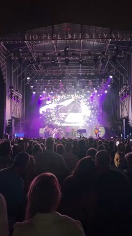 The video shows a wide shot of an outdoor concert stage at night. The stage is elaborately lit with purple and white lights, and a large screen displays abstract patterns. Several musicians are performing on stage, mostly visible as silhouettes against the bright lights. In front of the stage, a large crowd of people stands, facing the performers. Many in the crowd are holding up their phones, likely to record the event. The overall atmosphere appears to be energetic and celebratory, with the "TIO PEPE FESTIVAL" signage visible above the stage. The camera is positioned behind the crowd, looking towards the stage.
