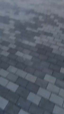 The video shows a paved driveway with grey and black interlocking stones. In the background, there is a neatly trimmed green hedge. To the left of the hedge, there is a small lawn with three decorative blue pots, two of which contain plants. Beyond the hedge, there is a red building with a dark roof and a dark-colored structure on the right side of the frame. In the distance, there are trees and mountains under a cloudy sky. The video is shaky and appears to be filmed from a moving vehicle or by someone walking. The lighting suggests it is either early morning or late evening.