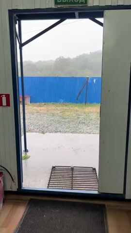 The video opens from inside a structure looking out through an open doorway. A metal grate is visible on the ground just outside the doorway. Beyond that, a gravel-covered area leads to a blue fence. On the other side of the fence, a wooded hillside is shrouded in a misty, rainy atmosphere. A small red-roofed structure, resembling a dog house, sits near the blue fence. The weather is overcast and raining, with visible raindrops falling. The camera pans slightly to the right, revealing more of the blue fence and the surrounding environment. The overall scene suggests an outdoor, possibly rural setting experiencing inclement weather.