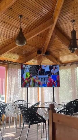 The video displays a television screen mounted on a wooden ceiling with exposed beams. The screen shows a group of male soccer players in red uniforms celebrating. They are hugging and appear to be in high spirits, suggesting a recent victory or significant achievement in a match. One player in the foreground is wearing a jersey with the number 13 and the name "RENATO VEIGA" visible on the back. Other players have numbers 7 and 10 on their jerseys. Behind the celebrating players, other individuals are visible, some in white uniforms. The setting appears to be an indoor viewing area, possibly a cafe or restaurant, indicated by the presence of tables and chairs in the foreground and decorative curtains in the background. The overall atmosphere conveyed is celebratory and exciting, likely during a sporting event. The lighting suggests it is daytime.