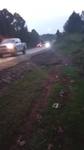 The video shows a view of a road at dusk or early evening. Cars are passing on the road. The road is lined with trees. The person recording is on the side of the road. They are speaking but the speech is not clear.