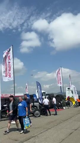 The video captures an outdoor event or exhibition under a bright, partly cloudy sky. Several men are seen walking and standing on a paved area, seemingly browsing the displays. In the background, a large white truck is visible alongside numerous large tires, suggesting the event's focus might be on agricultural equipment, heavy machinery, or related industries.

Adding an unexpected element to the scene, two helicopters can be observed flying in the distance against the blue sky.

Various flags are prominently displayed throughout the area. White flags feature Cyrillic text such as "МЕДИЦИНСКА ПОМОЩ" (Medical Assistance), "СТОПАНСКИ ДВОР" (Farm Yard/Agricultural Yard), "WWW.STOPANSKIDVOR.COM", and "МЕ-АНХИЕМ". Blue flags display "MICHELIN" with its well-known logo, and "CAT SPECIALTY" accompanied by the CAT logo. One man is carrying a bag with partially visible Cyrillic text, including "НАРЪЧНИК" (Handbook or Manual).

The event appears to be taking place in an open field or paved area, with hills or mountains visible in the background under the sunny conditions, consistent with the context of Madzherito, Bulgaria.