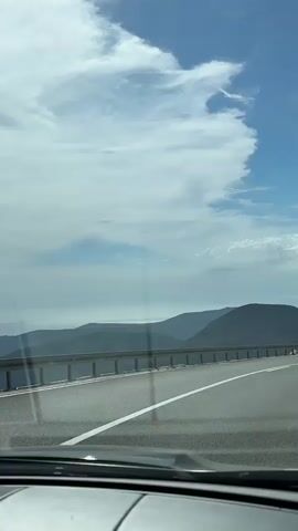 This video captures a view from inside a moving vehicle, likely a car, looking forward and slightly to the right through the windshield. The road ahead is paved and has a white line marking the lane. On the left side of the road, there's a guardrail overlooking a scenic landscape of rolling green hills and the sea in the distance. The sky is blue with scattered white clouds. Several cars are visible on the road ahead, also moving in the same direction. The interior of the vehicle is partially visible at the bottom of the frame, showing parts of the dashboard. The overall impression is of a scenic drive on a clear day.
