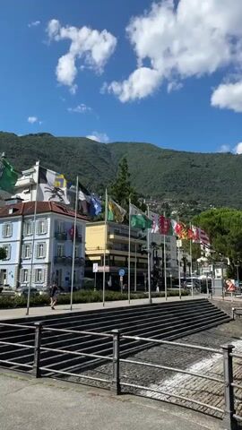 The video shows a scenic view of a waterfront with buildings and mountains in the background. A row of flags on poles stands along a promenade in front of the buildings. The flags are of various colors and designs, and some appear to be national flags. In front of the flags, there is a sidewalk and a railing overlooking a body of water. The water has a stone embankment. A person is walking on the sidewalk. The sky is blue with scattered white clouds, and lush green mountains rise behind the buildings. The overall impression is one of a pleasant day in a European town.