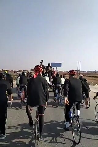 A group of cyclists rides down a road towards a small crowd standing next to a truck. Most of the cyclists wear black jackets and dark pants, with helmets of various colors. The truck appears to be carrying people with cameras, and there's a blue sign visible. The road is surrounded by a dry, dusty landscape with a few buildings in the distance. The sky is clear and blue. A couple of cyclists briefly embrace. The camera is positioned behind the cyclists, capturing their movement towards the crowd.