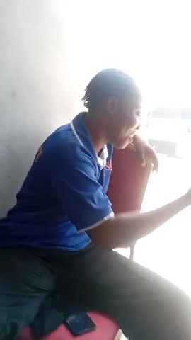 A woman with braided hair is sitting on a chair outdoors, leaning against a wall. She is wearing a blue polo shirt with writing on the back and black pants.  She is holding up a carton of coconut water. She looks to the side, then at the coconut water, and appears to be speaking. A cell phone lies on the seat beside her. The background shows a building and a bright, overexposed sky.