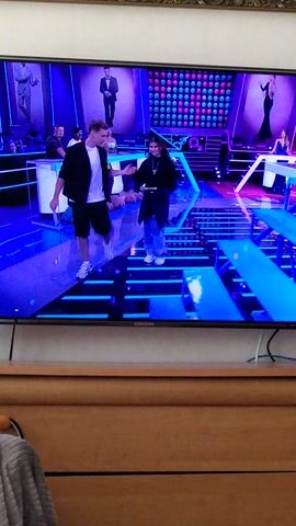 This media file shows a television screen displaying a scene with several people and a vibrant, colorful set. The primary subjects are two individuals prominently positioned on a platform in the foreground. The person on the left is a male with blonde hair, wearing a black jacket over a white shirt and dark shorts. He appears to be speaking or gesturing. To his right and slightly behind him is a female with dark hair, dressed in a black top and dark pants. She is also looking towards the front and seems to be interacting.

The setting is a brightly lit television studio with a stage featuring a multi-colored, patterned backdrop and blue, illuminated structures. In the background, there are several framed images or screens displaying silhouetted figures in various poses, some in dresses. The overall aesthetic is modern and energetic, with a strong emphasis on blue and pink lighting.

The activity appears to be a live broadcast or recording, possibly a game show, talk show, or performance, given the studio environment and the way the individuals are presented. The lighting suggests it could be either daytime or nighttime, as studio lights are used extensively. There are no clear weather cues.

Notable details include the Samsung logo visible on the right side of the TV screen, indicating the brand of the television. There are also some cables connected to the TV, one blue and one white, running vertically along the right edge of the frame. The people on screen appear engaged and alert, suggesting a professional broadcast environment. No specific text is legible on the screen itself.

The media file is oriented vertically, indicating it was likely filmed with a phone. The background of the room where the TV is located appears to be a wooden wall, suggesting an indoor setting.