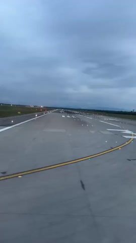 The video captures a ground-level perspective from an aircraft or vehicle moving along an airport surface at dusk or night. The scene is dominated by a concrete runway or taxiway, illuminated by numerous bright white, red, and green lights that line the path ahead and to the sides. The surface appears reflective, possibly indicating wet conditions or just the intensity of the lighting.

Visible on the concrete in the foreground is a large white painted number "3," likely a runway or taxiway designation. White and yellow painted lines also mark the concrete surface. In the distance, various red lights are visible, and faint outlines of mountains or hills can be seen against the cloudy, dim sky. The sound of an aircraft engine is audible, suggesting forward movement. No people are visible. The setting is consistent with an airport in Ferno, Italy.
