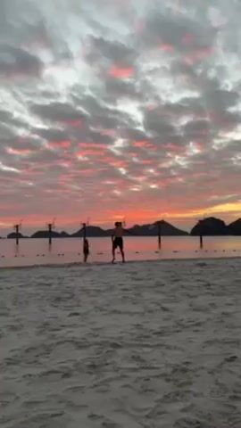 A man and a child stand on a sandy beach at dusk, their silhouettes visible against a vibrant sunset. The sky is filled with pink and gray clouds, casting a warm light across the scene. Several basketball hoops are visible in the water behind them, adding an interesting element to the landscape. A man walks across the beach in the foreground.