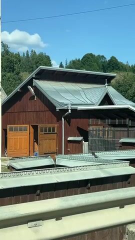 This is a video of a brown wooden building with a silver metal roof. The building appears to be a barn or workshop. There are two large wooden doors on the front of the building, one of which is partially open. There are also several smaller windows. In front of the building, there is a row of metal structures that look like either outdoor seating or storage containers. The background shows a green, tree-covered hillside under a clear blue sky with some scattered clouds. The video appears to have been filmed from a moving vehicle, as there is a blurred white barrier in the foreground.