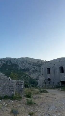 A vertical video pans across the ruins of a stone building. The building appears to be a fortification or old structure, with missing sections of wall and exposed openings that were once windows or doorways. The stonework is weathered and aged. The foreground consists of dry, rough ground with sparse vegetation, including dry grass and some small white flowers. In the background, a rocky, mountainous landscape rises under a clear, pale blue sky. The overall impression is one of an abandoned, historical site in a somewhat desolate natural setting.