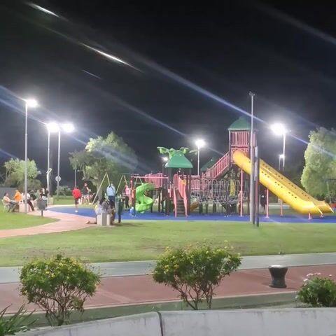At night, a brightly lit public park with play structures is visible. A playground with slides, climbing structures, and swings is centered in the frame, partially occupied by several people, including children playing and adults observing. Street lamps illuminate the area, casting long, stark lines of light. To the left, people are seated on benches. A paved path runs along the foreground, with small bushes separating it from the playground area. A cyclist rides on the path to the right.  The scene appears calm and populated with people enjoying the evening in the well-lit park.