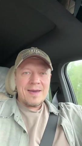 A man is sitting in the driver's seat of a car, wearing a light green cap and a light green button-down shirt over a beige t-shirt. He's got a seatbelt on. He appears to be singing, or at least speaking and emoting. The interior of the car is black and beige. Through the window, trees can be seen passing by.