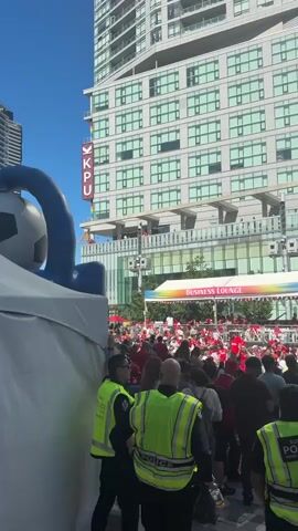 The video shows a crowd of people gathered outside a modern building. In the foreground, police officers wearing high-visibility vests are present. To the left, a large inflatable soccer ball is partially visible, along with a white tent. A banner for a "BUSINESS LOUNGE" is visible in the middle ground. The building itself has multiple floors with large windows, and a sign with the letters "KPU" and a rainbow flag is displayed on its facade. People are seen on balconies and walkways on the building's upper levels, some wearing red shirts. The overall scene suggests an outdoor event or gathering.