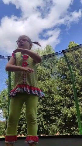A young girl with blonde pigtails and glasses is jumping on a trampoline. She is wearing a lime green shirt with red and pink flower accents and lime green leggings with red and pink ruffles at the bottom. The trampoline is surrounded by a green safety net, and lush green trees and a blue sky with white clouds are visible in the background. The girl appears to be mid-air, with her arms outstretched and her body angled as if she is performing a jump or trick.