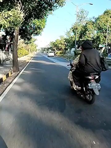 A person wearing a dark hoodie rides a scooter on a paved road. The road is lined with trees and a curb on the left side. Another scooter is visible further down the road. The sky is clear and blue.