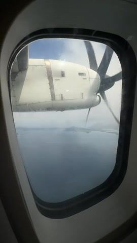A view from inside an airplane window shows a white aircraft engine with a rotating propeller against a backdrop of cloudy sky and a distant horizon of water and land. The window frame is partially visible. The camera is steady.