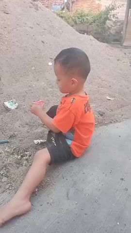 A young boy wearing an orange t-shirt and black shorts sits barefoot on a gray concrete surface. He is holding a small yellow cup and appears to be playing with sand, pouring it from the cup onto his hand. In the background, a large pile of sand or dirt is visible, along with some debris on the ground and houses in the distance. The boy looks down at his hands as he plays.
