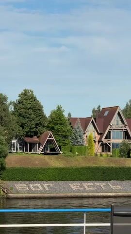 The video shows a large, multi-story house with a red roof and large windows, nestled amongst trees. In the foreground, a body of water is visible, with the railing of a boat in the lower right corner. Along the edge of the water, a line of stones forms the words "БОГ ЕСТЬ ЛЮБОВЬ" which translates to "God is Love". The sky is blue with some clouds.