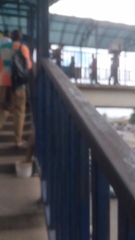 The video shows a group of people, seemingly waiting or resting, on a set of outdoor stairs. The perspective is from a low angle, looking up towards a metal structure with blue slats that runs along the top of the stairs. A portion of a person's leg, wearing a blue and white patterned garment, is visible in the foreground. In the background, several individuals are seated or standing on the steps. Some are wearing traditional clothing, while others have bags or backpacks. The overall environment appears to be an urban or semi-urban setting with a covered walkway overhead, suggesting a public space like a bus station or market. The lighting indicates daytime. The footage is shaky and the focus shifts intermittently.
