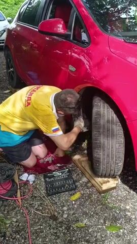 A man in a yellow shirt and blue shorts kneels on the ground next to a red car. He is working on the front passenger-side wheel, which is elevated on wooden blocks. A red cloth is spread out on the ground beneath him, with a set of tools on top. The man appears to be using a wrench to tighten or loosen something on the wheel assembly. The car is parked on a gravelly surface, with some green leaves scattered around. In the background, another white car is partially visible. The man has a Russian flag patch on his shirt.