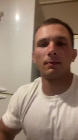 A man in a white t-shirt flexes his bicep in front of a refrigerator. He is looking at the camera. The video is dimly lit.