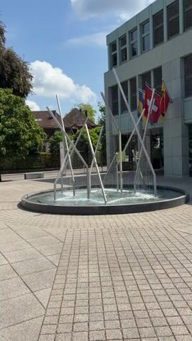 This video shows a circular fountain in front of a modern building. The fountain features several metallic poles of varying heights, with water spraying from the top of some of them, creating a cascading effect into the basin. The basin is surrounded by a patterned stone walkway. Behind the fountain, the building has a light-colored facade and several windows. Three flags are visible hanging from the building: a Swiss flag, and two other flags that are partially obscured but appear to be red and yellow. To the left of the building, there are trees and a hint of residential rooftops. The sky is blue with scattered white clouds. The overall scene suggests an outdoor public space on a sunny day.