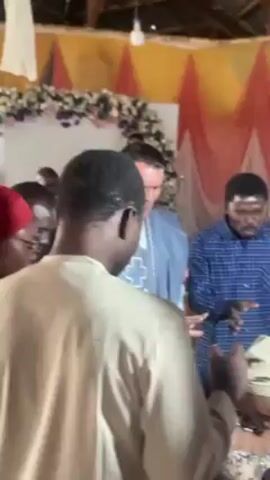 The video shows a group of men standing around a person sitting on a chair. The seated person is wearing a white head covering. The men are dressed in a variety of clothing, including traditional African attire and Western-style shirts. The man in the blue patterned shirt is gesturing with his hands while talking. The environment appears to be indoors, with a backdrop of colorful drapery and floral arrangements.