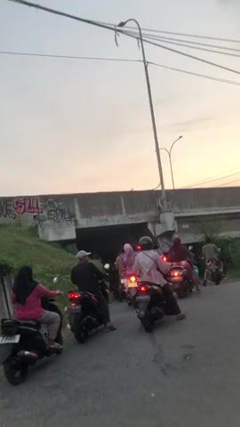 The video shows a group of people on motorcycles stopped at a road intersection. The time appears to be dusk, with a hazy sky. In the background, there is a concrete bridge with graffiti on its underside. Several motorcycles are visible, some with riders wearing headscarves. The motorcycles are facing forward as if waiting for a signal or traffic to clear. There are power lines overhead and some vegetation along the side of the road. The overall scene suggests a common urban or semi-urban setting.