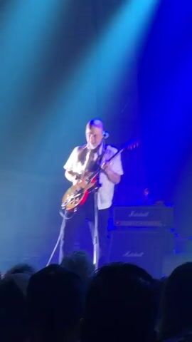 Here is a description of the video.
The video shows a person performing at a concert. The performer is a man who is bald. He is wearing a white, short-sleeved shirt and blue pants. He is standing on a stage, playing an electric guitar. Behind him are two Marshall amplifier stacks. The stage is lit with blue spotlights. In the foreground, there is an audience with their backs to the camera. One person is holding up the "rock on" hand gesture.