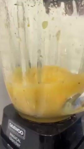 A Daewoo Magical blender is actively blending a yellow liquid with small dark pieces. The blender is partially translucent and shows water droplets. The liquid is being thoroughly processed within the blender's container. The blender's base is black.