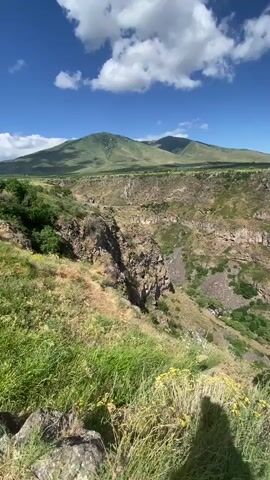 This media file captures a scenic vista of a deep canyon under a bright, blue sky dotted with fluffy white clouds. The foreground is dominated by lush green grass and rocky terrain. As the camera pans, the vastness of the canyon unfolds, revealing layered rock formations and vegetation clinging to the steep cliffs. 

In the distance, rolling hills and a flat, cultivated landscape stretch towards the horizon. A small cluster of buildings can be seen on a distant hilltop. Further along the canyon rim, a dirt path winds its way, and several people are visible enjoying the view. Some are standing near the edge, while others are walking along the path. Their presence adds a sense of scale to the immense landscape.

The bright sunlight and clear skies suggest a warm, pleasant day, likely in the late morning or early afternoon. The scene evokes a feeling of awe and tranquility, highlighting the natural beauty of the location. The time of day is consistent across the frames, with the sun casting shadows that indicate a high sun angle. 

While no visible text is present in the media, the description of the location as Artashavan, Armenia, provides geographical context, suggesting this is likely a well-known natural landmark in the region. The overall impression is one of a breathtaking natural landscape being appreciated by visitors on a beautiful day.