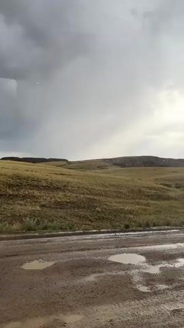 This is a video of a vast, open landscape in Karagayl, Kazakhstan. The scene is dominated by rolling hills covered in dry, golden-brown grass. In the foreground, a muddy, unpaved road is visible, with several puddles reflecting the overcast sky. The sky itself is a mixture of dark, heavy clouds and patches of brighter, diffused sunlight, suggesting an approaching storm or recent rainfall. The overall impression is one of a rugged, natural environment. There are no people or man-made objects visible other than the road itself. The time of day is not precisely clear but appears to be late afternoon or early evening, given the muted light. The weather is cloudy and possibly damp. The vastness of the landscape and the muted colors evoke a sense of solitude and perhaps a touch of melancholy due to the weather. There is no visible text in the video.