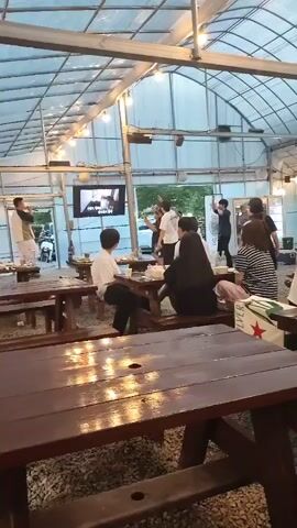 A group of people are gathered in a greenhouse-like structure with a clear plastic roof and metal supports. The space appears to be an outdoor dining or event area, with wooden picnic tables and benches. In the background, a television screen displays an image. A man on the left is standing and appears to be speaking or performing. Several people are seated at tables, some facing away from the camera, and a group of individuals are standing and interacting at the back of the space, where a performance or activity is taking place. The lighting suggests it is daytime, possibly late afternoon or early evening.