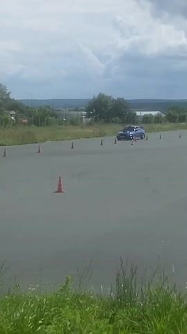 A blue Subaru Impreza is seen on a grey asphalt surface, likely a track or training area. The car appears to be in motion, possibly performing a maneuver as evidenced by the tire marks on the ground. Several orange traffic cones are strategically placed around the area, suggesting a course or obstacle setup. In the background, there is a green building, a line of trees, and a cloudy sky. The foreground is filled with tall green grass. The video seems to capture a segment of a driving event or practice session.