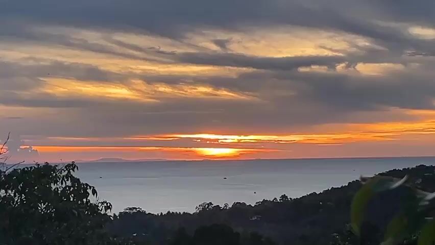 The video shows a scenic sunset over the ocean. The sky is filled with gray clouds streaked with orange and yellow from the setting sun. The sun is visible as a bright orange orb just above the horizon. The water is calm and reflects the colors of the sky. In the foreground, there is a treeline with green leaves. The scene is peaceful and serene.