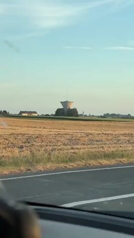 The video shows a view from a vehicle, likely a car, traveling on a road. The road is visible in the foreground, with a light-colored line marking its edge. Beyond the road, there is a field of dry, golden-colored crops, suggesting it is late summer or early autumn. In the middle ground, a distinctive water tower with a conical top stands prominently, surrounded by green trees. To the left of the water tower, a white house with a dark roof is visible. In the distance, more trees and some smaller structures, possibly other buildings or windmills, can be seen on the horizon. The sky is a clear, pale blue with a few wispy clouds. The overall impression is a rural landscape during daytime. The camera appears to be slightly tilted, and there are some smudges or specks on the windshield, which is also visible in the foreground, obscuring a portion of the view. The video is short, lasting only a few seconds, and seems to capture a fleeting moment of travel through the countryside.